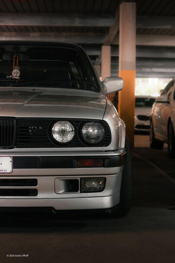 A silver BMW E30 in a parking garage, lit by the morning light